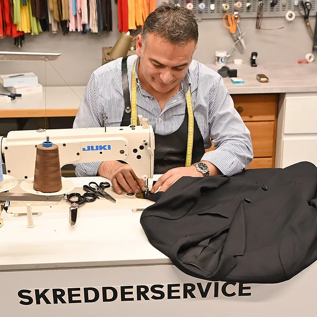 Skredder syr en svart dressjakke på en industriell symaskin i skredderverkstedet.